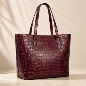 May include: A deep burgundy tote bag featuring a textured crocodile-embossed pattern. The bag has a structured rectangular shape with two vertical handles attached to the top edge, providing a clean and functional design for carrying items.