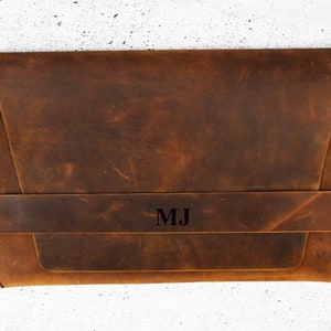 May include: A brown leather laptop sleeve with a flap closure and a strap with the initials "MJ" embossed on it.
