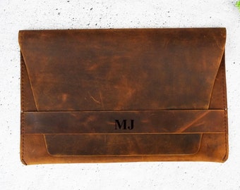 May include: A brown leather laptop sleeve with a flap closure and a strap with the initials "MJ" embossed on it.