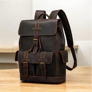 May include: A dark brown leather backpack with multiple pockets and adjustable straps. The backpack features a flap closure with buckles and a top handle. The stitching is a contrasting orange color, and the bag appears to be made of high-quality leather.