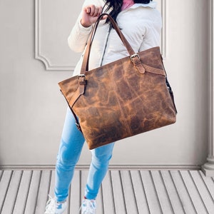 May include: A brown leather tote bag with a distressed finish. The bag has two top handles and adjustable shoulder straps. It features metal buckles and a rectangular shape. The bag is held by a person wearing a white jacket and blue jeans.