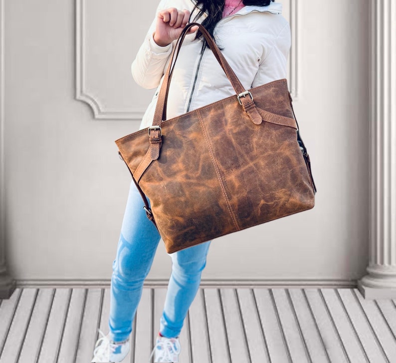 May include: A brown leather tote bag with a distressed finish. The bag has two top handles and adjustable shoulder straps. It features metal buckles and a rectangular shape. The bag is held by a person wearing a white jacket and blue jeans.