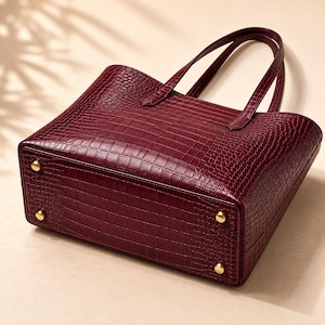 May include: A deep burgundy tote bag featuring a textured crocodile skin pattern. The bag has two thin handles and four small, round, gold-toned metal feet attached to the rectangular base, providing stability when the bag is placed on a flat surface.