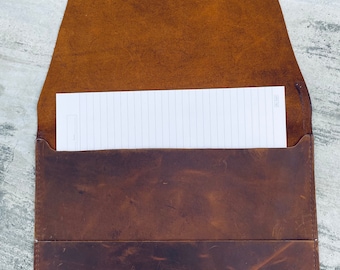May include: A brown leather folder with a flap closure and a lined notepad inside. The folder has a distressed look and a brown leather strap.