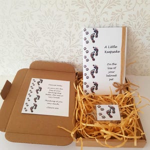 Puede incluir: Una caja de cartón marrón que contiene un libro de recuerdos blanco, una tarjeta y un pequeño llavero. El libro y la tarjeta presentan diseños de huellas de patas y el texto "A Little Keepsake" y "On the loss of your beloved pet."