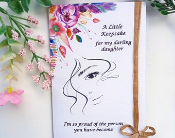 May include: A white card with a watercolor floral design and the text "A Little Keepsake for my darling daughter I'm so proud of the person you have become". The card is tied with a brown ribbon.