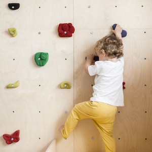 Könnte beinhalten: Ein Kind klettert an einer Holzkletterwand mit bunten Griffen. Die Wand ist aus Sperrholz und hat eine Vielzahl von Formen und Größen an Griffen. Das Kind trägt ein weißes Hemd und eine gelbe Hose.