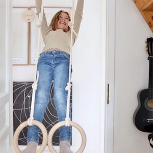 May include: A child is hanging from wooden gymnastic rings suspended from a wooden bar in a doorway. The rings are attached to the bar with white ropes. The person is wearing a light-coloured long-sleeve top and blue jeans. The rings are for indoor exercise.