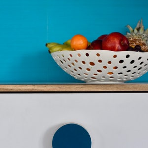 May include: A white ceramic fruit bowl with a variety of fruit, including bananas, apples, and a pineapple, sits on a light wood countertop. The bowl has a unique design with small holes throughout. A blue round knob is on the white cabinet door below the bowl.