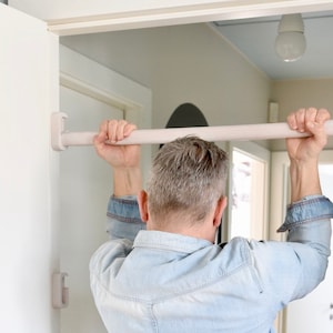 Könnte beinhalten: Eine Person verwendet eine weiße Spannstange, um etwas an einem Türrahmen aufzuhängen. Die Stange ist mit zwei weißen Kunststoffhalterungen am Türrahmen befestigt.