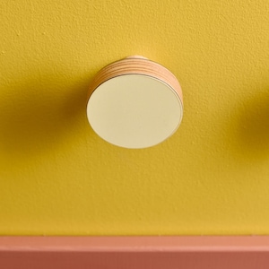 Könnte beinhalten: Eine runde, hölzerne Deckenleuchte mit einem weißen, runden Schirm. Die Leuchte ist an einer gelben Wand mit einer braunen Holzverkleidung darunter montiert.