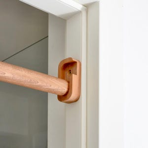 May include: A wooden curtain rod bracket mounted to a white door frame. The bracket is made of natural wood and has a rounded shape. The rod is also made of wood and is attached to the bracket.