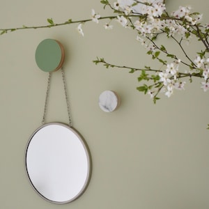 Puede incluir: Un espejo redondo con un marco plateado cuelga de una cadena unida a un pomo de madera verde. Dos pomos redondos, uno verde y uno blanco, están montados en la pared. Una rama florida con flores blancas está en el fondo.