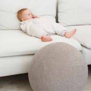 Könnte beinhalten: Ein Baby in einem weißen Strampler liegt auf einem weißen Sofa mit einem grauen, strukturierten Ball im Vordergrund.