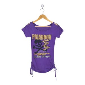 May include: A purple t-shirt with a scoop neck and short sleeves, hanging on a wooden hanger. The shirt features the text "PICAROON" and a skull and crossbones graphic. The text "COMBAT ANY KIND OF RIOTING" is also visible.