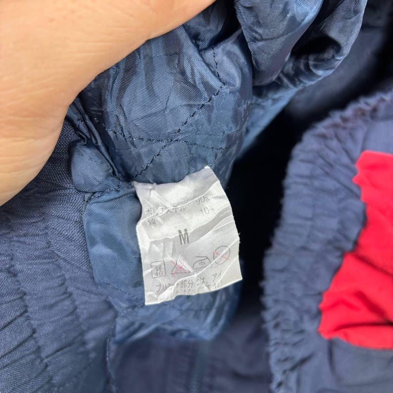 May include: Close-up of a navy blue garment with a red accent. A white tag with Japanese text and the letter "M" is visible. The garment appears to be made of a quilted material.