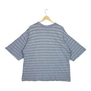 May include: A gray and blue striped t-shirt with short sleeves hangs on a wooden hanger. The shirt has horizontal stripes in varying shades of gray and blue. The neckline is a solid blue color.