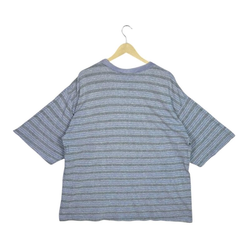 May include: A gray and blue striped t-shirt with short sleeves hangs on a wooden hanger. The shirt has horizontal stripes in varying shades of gray and blue. The neckline is a solid blue color.