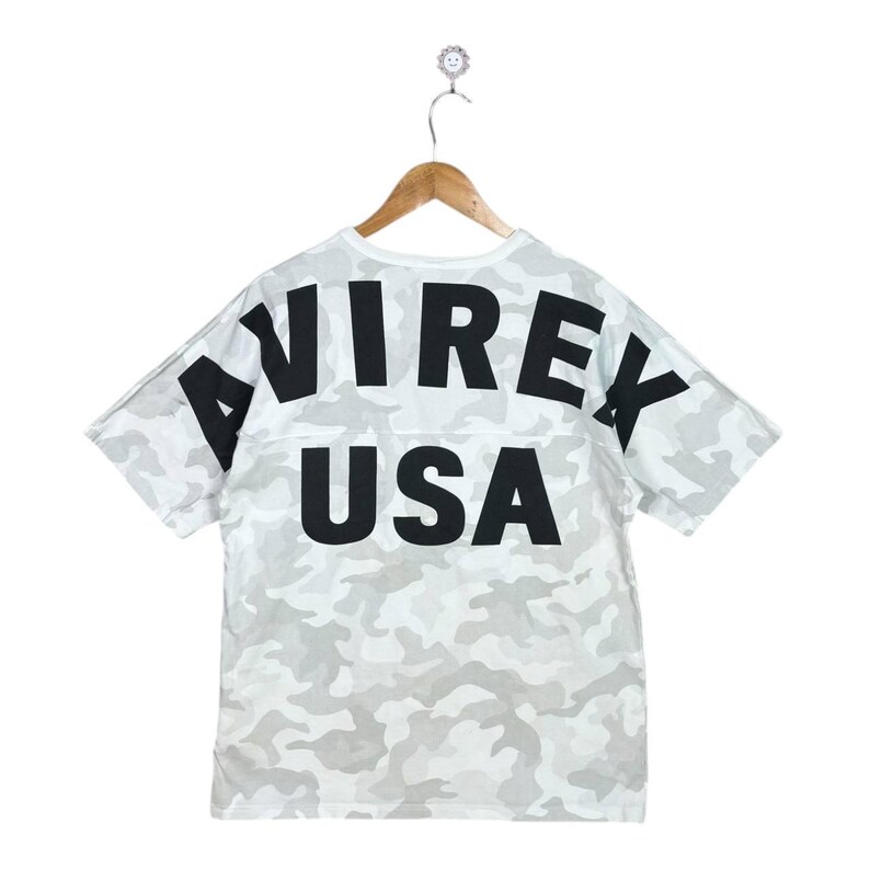 May include: A short-sleeve t-shirt featuring a light grey and white camouflage pattern. The back of the shirt displays the text AVIREX in a large, bold, black arched font, with the word USA printed in a large, bold, black font directly below it.