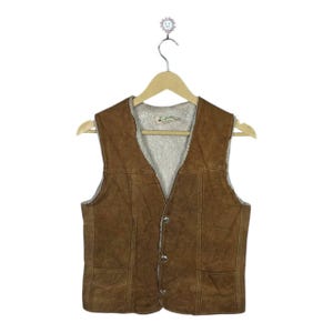 May include: Brown suede vest with a cream-colored, fuzzy lining. The vest has a V-neck, two front pockets, and three buttons down the front. It is hanging on a wooden hanger.