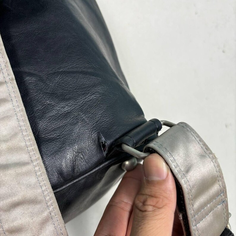 Op de afbeelding: Close-up van een donkerblauwe leren tas met een zilveren riem en hardware. De tas heeft een getextureerd oppervlak en een zichtbare scheur bij de riembevestiging. Een lichtbeige stof is zichtbaar aan de zijkant.