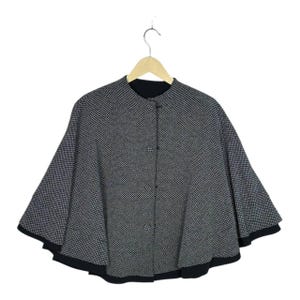May include: A black and white houndstooth patterned cape with a round neckline and a front button closure. The garment features a flared silhouette and a solid black hemline along the bottom edge, displayed on a wooden hanger against a white background.