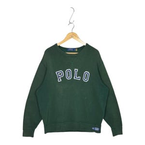 May include: A dark green long-sleeve sweatshirt featuring the word POLO in large, arched, navy blue letters with white outlines across the chest. The garment has a crew neckline with a V-stitch detail and a small rectangular label at the hem.