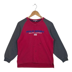 May include: A vintage red and gray Polo Sport sweatshirt with a V-neck. The sweatshirt features the Polo Sport logo in blue, the Ralph Lauren text, and a small American flag. The sleeves are gray, and the body is red.