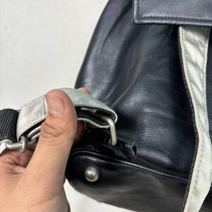 Op de afbeelding: Close-up van een zwarte leren rugzak met een zilveren band en accenten. De band is met een zilveren metalen sluiting aan de tas bevestigd. De tas heeft een kleine scheur aan de onderkant.