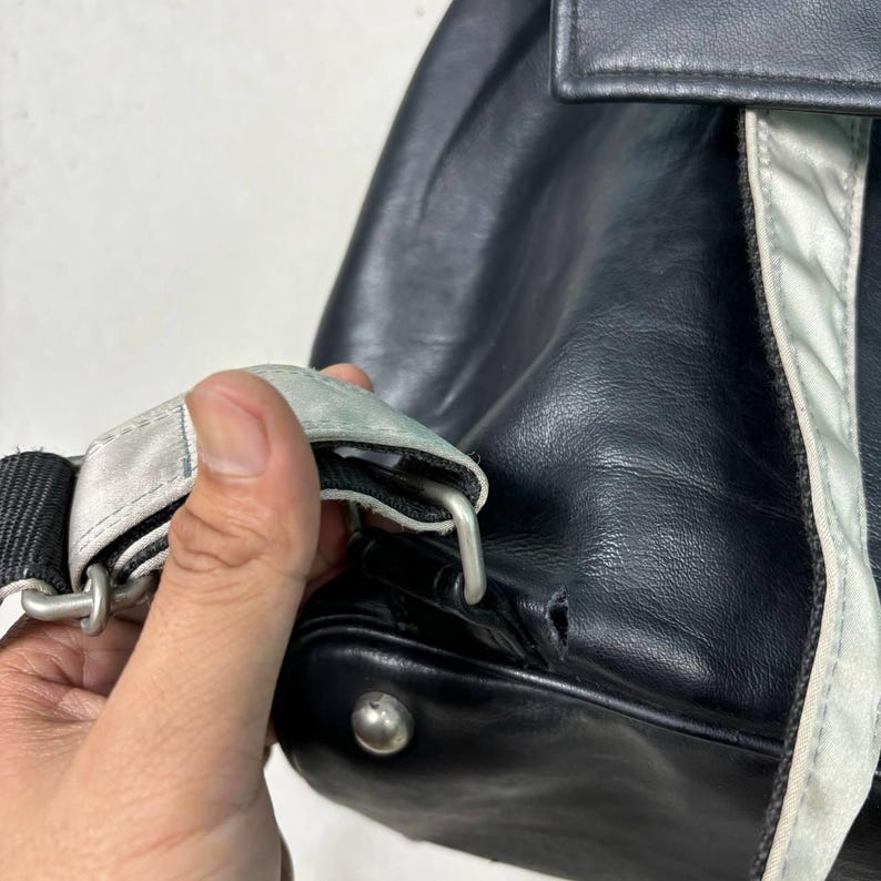 Op de afbeelding: Close-up van een zwarte leren rugzak met een zilveren band en accenten. De band is met een zilveren metalen sluiting aan de tas bevestigd. De tas heeft een kleine scheur aan de onderkant.