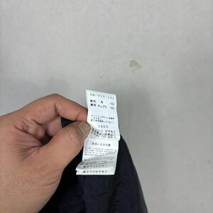 Op de afbeelding: Een hand die een wit kledinglabel met zwarte tekst vasthoudt. Het label toont het stijlnummer "YK-P15-102" en stofdetails. Japanse tekst en wasvoorschriften zijn ook zichtbaar. De stof lijkt donker.