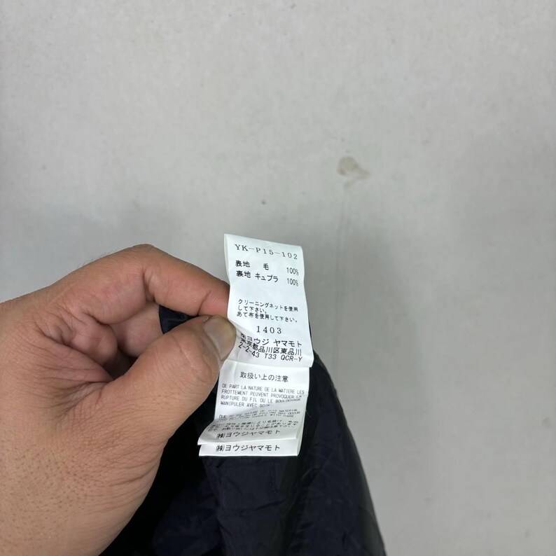Op de afbeelding: Een hand die een wit kledinglabel met zwarte tekst vasthoudt. Het label toont het stijlnummer "YK-P15-102" en stofdetails. Japanse tekst en wasvoorschriften zijn ook zichtbaar. De stof lijkt donker.