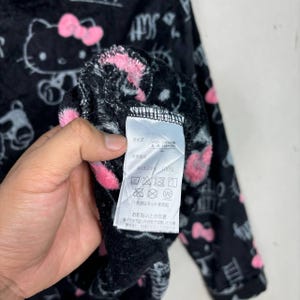 Puede incluir: Tela de felpa negra con un patr&oacute;n repetido de Hello Kitty y lazos rosas. Una mano sostiene la tela y una etiqueta con texto japon&eacute;s es visible. La tela tiene una textura suave y difusa.