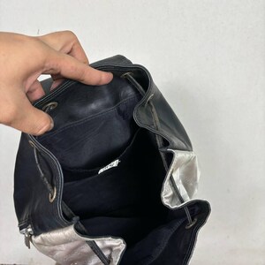 Op de afbeelding: Zwarte leren rugzak met een zilverkleurig accent. De tas heeft een trekkoordsluiting en een ritsvak. De binnenkant is zwart en de tas lijkt open te zijn, waardoor de binnenkant zichtbaar is.