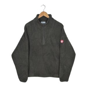 May include: A dark olive green fleece pullover with a half-zip closure and a small logo patch on the upper left sleeve. The fleece is hanging on a wooden hanger against a white background.