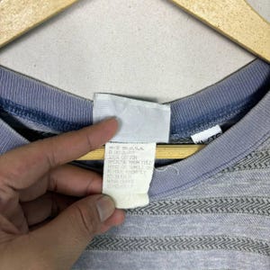 May include: Close-up of a gray and blue Quiksilver t-shirt. The shirt has a striped pattern across the chest and a blue collar. The tag reads "MADE IN U.S.A. QUIKSILVER 100% COTTON".
