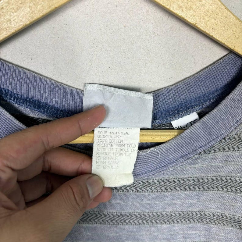 May include: Close-up of a gray and blue Quiksilver t-shirt. The shirt has a striped pattern across the chest and a blue collar. The tag reads "MADE IN U.S.A. QUIKSILVER 100% COTTON".