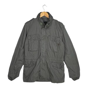 May include: An olive green military-style jacket with multiple front pockets and a high collar. The jacket is suspended from a wooden hanger. The jacket has a button closure and epaulettes on the shoulders.