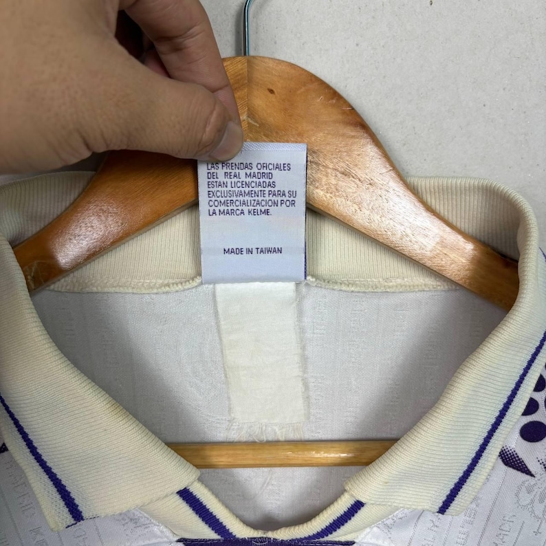K&ouml;nnte beinhalten: Cremefarbenes Trikot mit lila und wei&szlig;em Design. Der Kragen und die &Auml;rmel haben lila Streifen. Das Etikett lautet "LAS PRENDAS OFICIALES DEL REAL MADRID ESTAN LICENCIADAS EXCLUSIVAMENTE PARA SU COMERCIALIZACION POR LA MARCA KELME MADE IN TAIWAN."