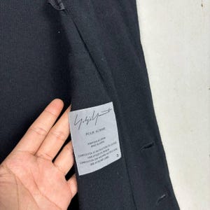 Może przedstawiać: Czarny płaszcz z etykietą z napisem "Yohji Yamamoto POUR HOMME". Etykieta zawiera r&oacute;wnież tekst w języku francuskim i angielskim. Płaszcz ma trzy guziki i teksturowaną tkaninę.
