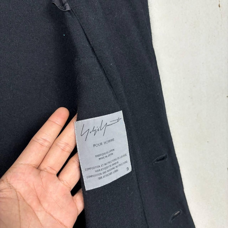 Może przedstawiać: Czarny płaszcz z etykietą z napisem "Yohji Yamamoto POUR HOMME". Etykieta zawiera r&oacute;wnież tekst w języku francuskim i angielskim. Płaszcz ma trzy guziki i teksturowaną tkaninę.