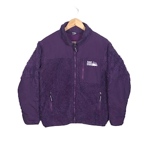 Puede incluir: Una chaqueta de forro polar de color morado con cierre de cremallera y dos bolsillos. La chaqueta tiene el texto "FIRST DOWN" y un logotipo de montaña en la parte delantera.
