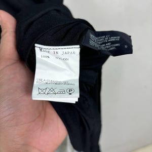 May include: Close-up of a black garment with a white tag. The tag reads "MADE IN JAPAN" and "100% NYLON." Washing instructions are also visible. The garment is held up against a light background.