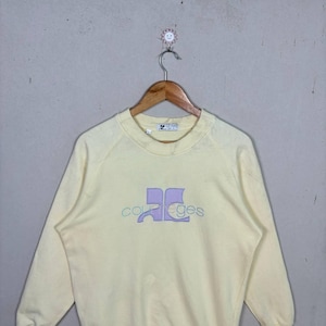 Pode incluir: Uma camisola amarela p&aacute;lida com gola redonda e mangas compridas. A frente apresenta o nome da marca "Courreges" num design lavanda e azul claro. A camisola est&aacute; pendurada num cabide de madeira.