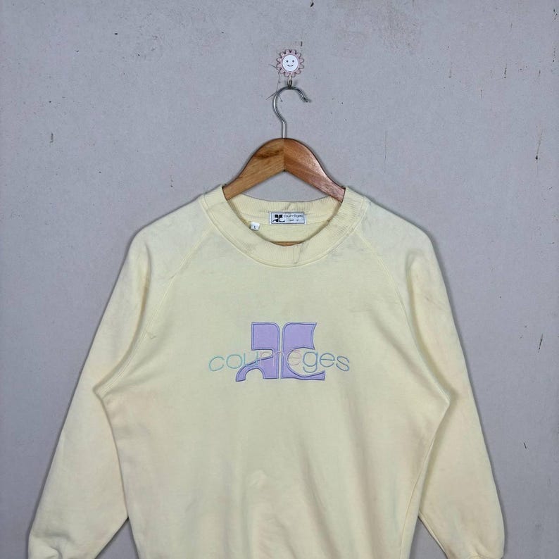 Pode incluir: Uma camisola amarela p&aacute;lida com gola redonda e mangas compridas. A frente apresenta o nome da marca "Courreges" num design lavanda e azul claro. A camisola est&aacute; pendurada num cabide de madeira.