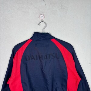 May include: Navy blue zip-up jacket with red side panels. The word "DAIHATSU" is printed across the back in black. The jacket is hanging on a metal hanger against a gray background.
