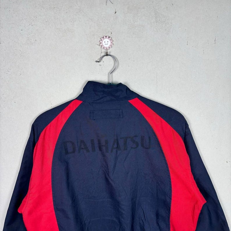 May include: Navy blue zip-up jacket with red side panels. The word "DAIHATSU" is printed across the back in black. The jacket is hanging on a metal hanger against a gray background.