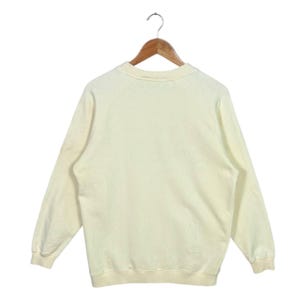 Pode incluir: Um moletom creme com mangas compridas, pendurado em um cabide de madeira. O moletom tem gola redonda e punhos e bainha canelados. O tecido parece ser um material macio e confort&aacute;vel.