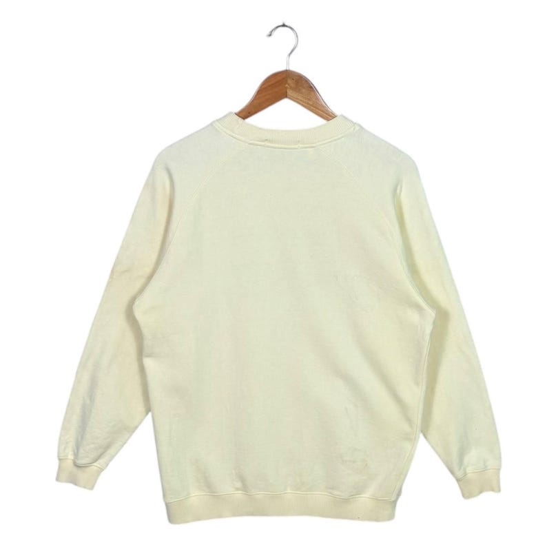 Pode incluir: Um moletom creme com mangas compridas, pendurado em um cabide de madeira. O moletom tem gola redonda e punhos e bainha canelados. O tecido parece ser um material macio e confort&aacute;vel.