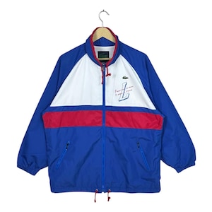 May include: A blue, white, and red Lacoste windbreaker jacket. The jacket features a full zip, a white panel across the chest, and a red stripe. The Lacoste logo is on the chest, and the text "I love tennis" is printed on the right side.